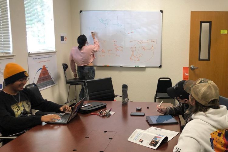 students sit at a table while one student writes on a white board