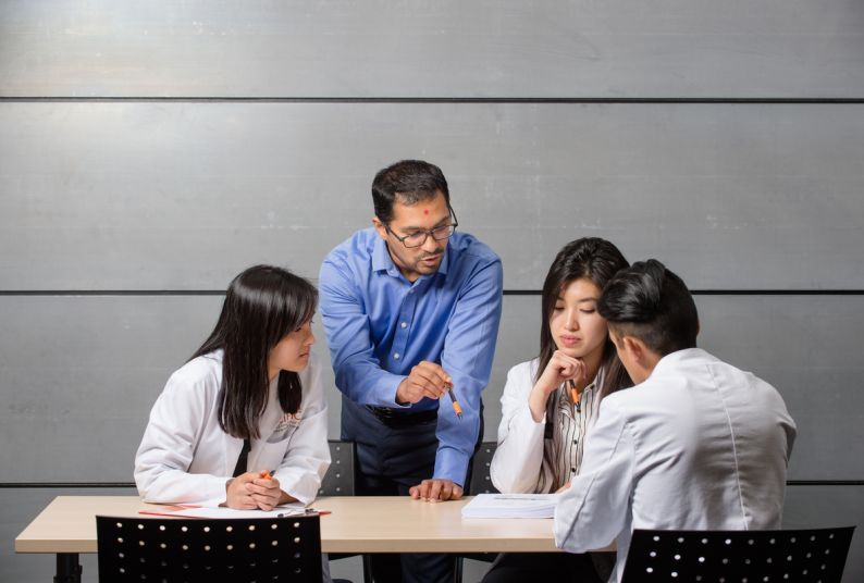 a professor stands next to a table with three seated students